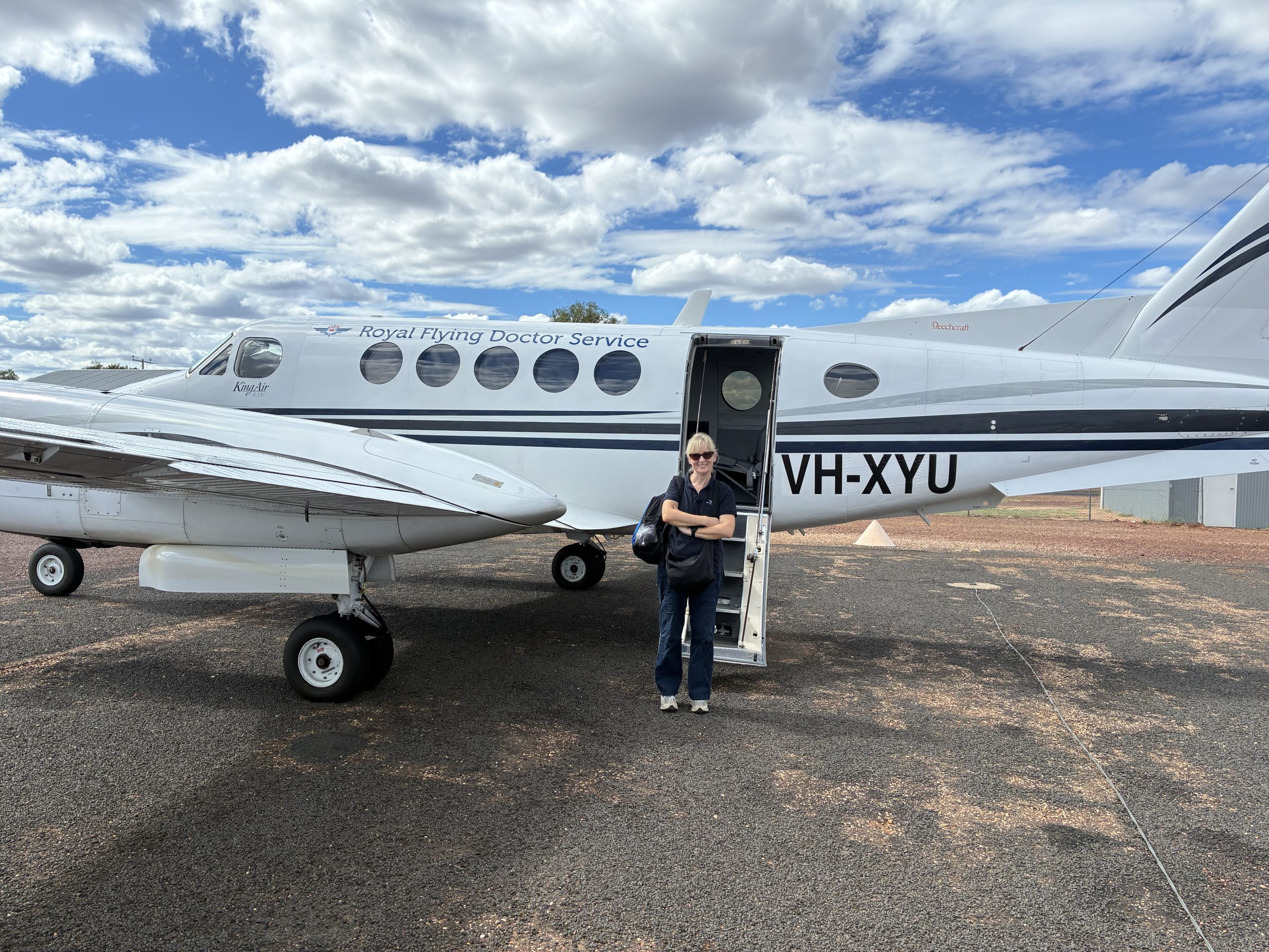 A woman wearing dark blue scrubs stands in front of a Royal Flying Doctor Service small plane on a regional airstrip. She smiles at the camera