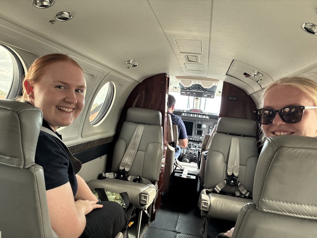 Two young women sit inside a Royal Flying Doctor Service plane, you can see the pilot at the front of the cabin. They are part of the dental team.